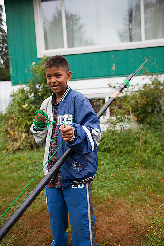 A boy stands in front of a house, holding onto a spear. The spear is taller than the boy. A green rope attached to the spear is wrapped around the boy’s hand.