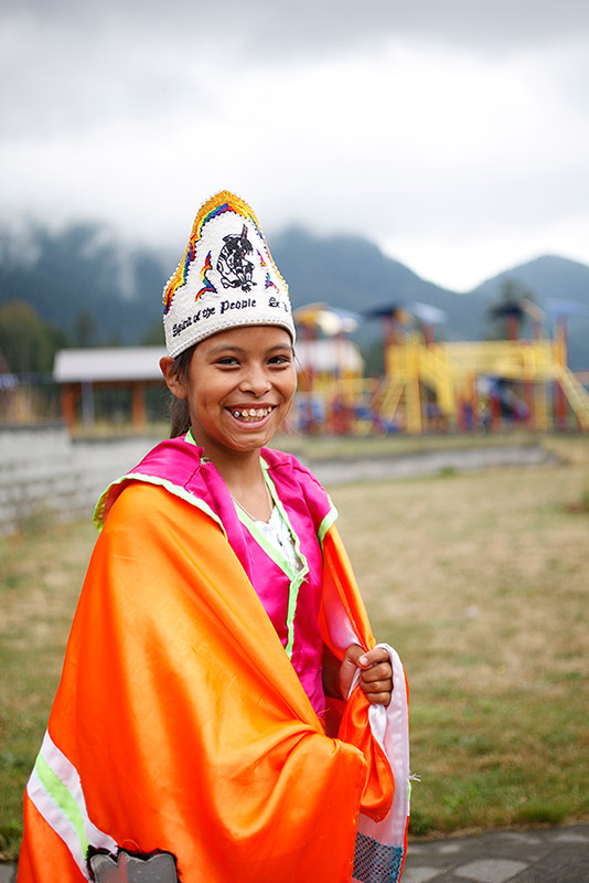 A girl wearing a fancy powwow dress smiles at the camera. The dress is bright orange, pink, and white. Her hat is white and has a black design on it.