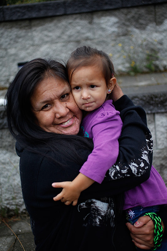 A woman holds her young granddaughter in her arms. The woman is smiling. Her granddaughter is wearing a purple dress and stares into the distance.