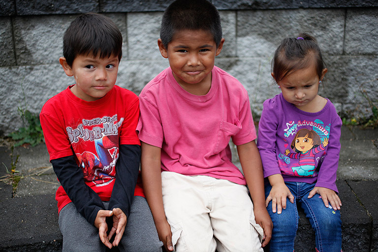 Two boys and a girl sit in front of a grey stone wall. The boys are smiling and the little girl looks pensive.
