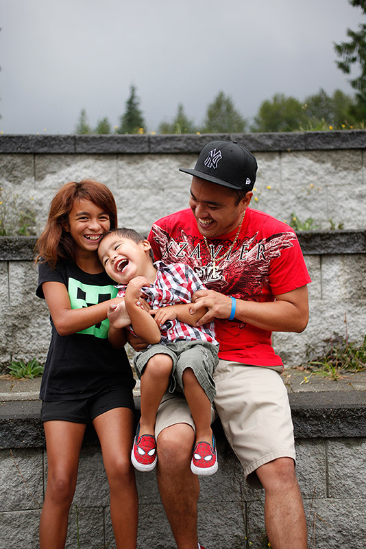 A girl, a boy, and a man sit in front of a grey stone wall. All three are laughing. The boy sits on the leg of the man.