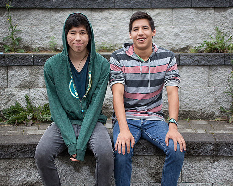 Two teenage boys sit in front of a grey stone wall. They are both smiling at the camera.