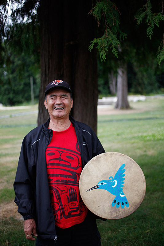 Un homme debout devant un cèdre et il rit. Il tient un tambour avec un dessin de colibri bleu.