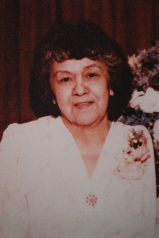 An older photograph of a woman standing in front of a red curtain. She is wearing a fancy white dress that has a floral brooch pinned on it.