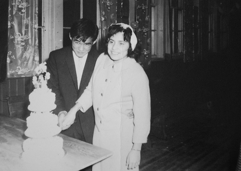 Une photo en noir et blanc d’un jeune homme et d’une jeune femme le jour de leur mariage. Ils coupent le gâteau de noces.