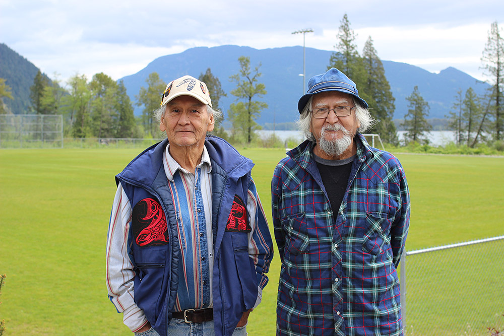 Deux hommes debout devant un terrain de soccer. À gauche, l’homme porte une veste bleue avec des motifs rouges. À droite, l’homme a les mains derrière le dos et il porte une chemise bleue à carreau.