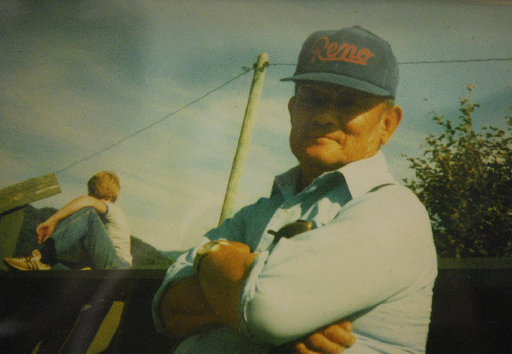 A man stares into the camera with his arms crossed. He is wearing a blue shirt and a blue baseball cap that has the word “Reno” printed on it in red.