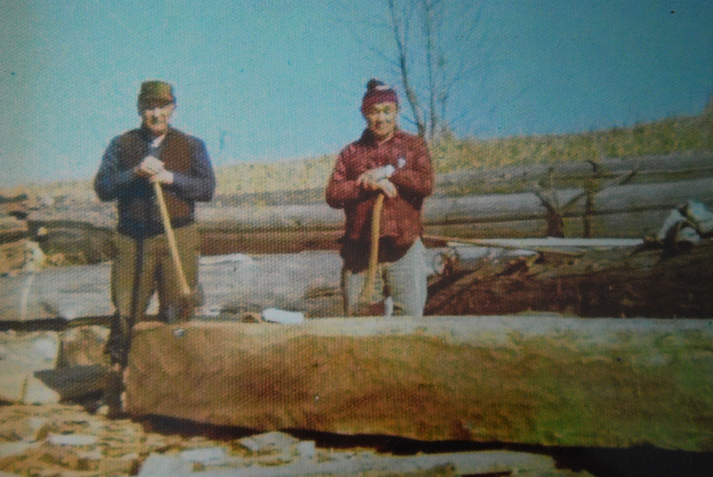 Deux hommes debout derrière un billot. Chacun tient une hache qui repose sur le billot.