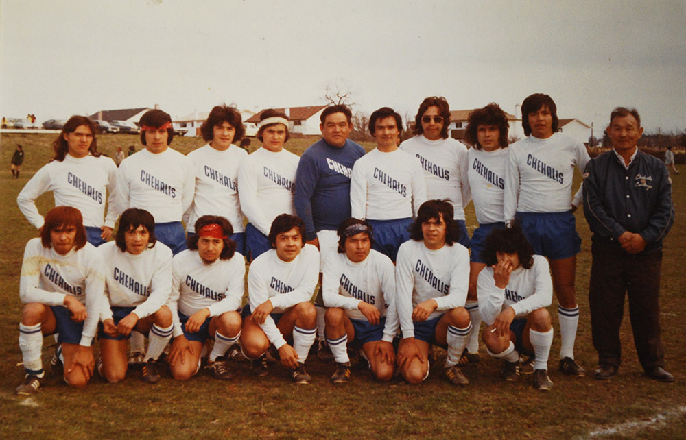 A soccer team poses for a photo. All the men except two are wearing white soccer jerseys, blue shorts, and white socks.