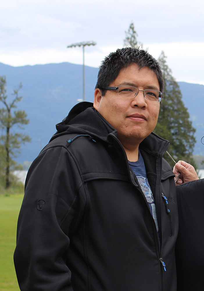 A man stands in front of a soccer field. In the background are some trees, a body of water, and mountains. He is staring into the camera.