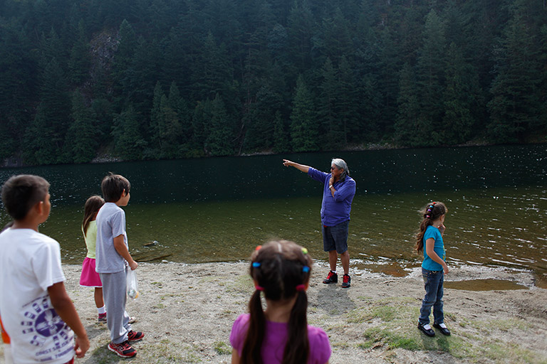 A man on the shoreline points to a place off-camera. Children are listening in the foreground.