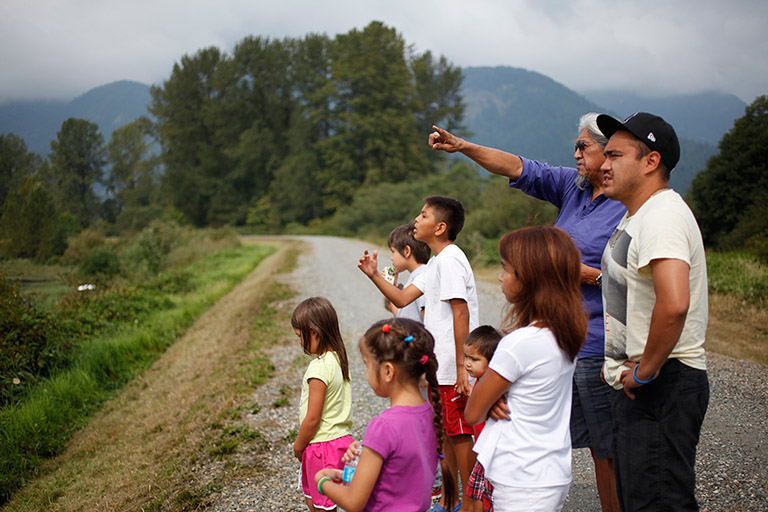 Two men and a group of children stand at the side of a road. They are staring to the side of the road at one of the men is pointing at.