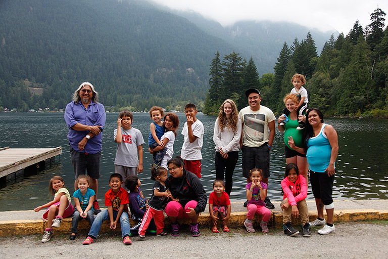A group of children and young adults pose with their elder for a photograph on the shore of the river. There are mountains and trees in the background.
