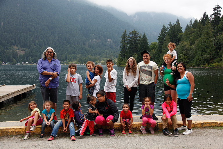 Un groupe d’enfants et de jeunes adultes posent avec leur aîné pour une photo sur le rivage. À l’arrière-plan, il y a des montagnes et des arbres.