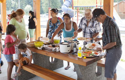 Children, youth  and adults, both male and female, are gathered around a picnic table serving food.