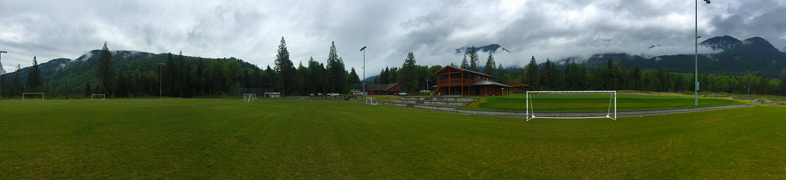 A soccer pitch is in the foreground of the image, with trees and mountains in the background. The sky above is grey and cloudy.