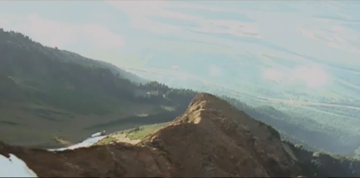 An aerial photograph shows the peak of a mountain, mainly brown with some forested areas below and in the background.