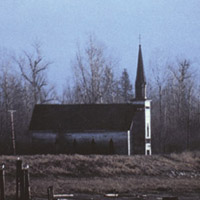 A side view of a small church in a field. In the background is a forest and a snow-covered mountain.