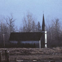 Une vue de côté d’une petite église dans un champ. À l’arrière-plan, une forêt et une montagne au sommet enneigé.