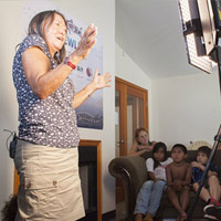 A woman is standing on the left side of the photo, sharing a story with children that are seated on a couch in the background. There is a woman on the right side that is filming the event.